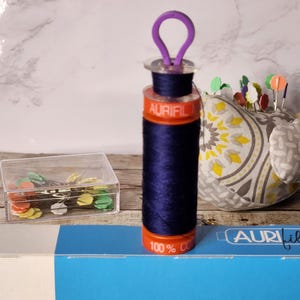 May include: A spool of dark blue thread with a purple plastic thread holder. The spool is labeled "Aurifil" and "100% Cotton".