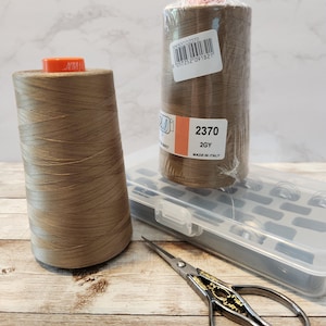 May include: Two spools of beige thread, one with a label that reads "2370 2GY MADE IN ITALY", a clear plastic box with compartments, and a pair of small silver scissors with gold accents.