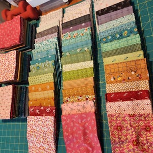 May include: Stacks of colorful fabric squares in various patterns and colors, including pink, green, orange, and blue. The fabric is arranged on a green cutting mat, suggesting a quilting or sewing project. Scissors and a small red weight are visible.