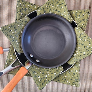 May include: A black frying pan with an orange handle rests on a green floral patterned pot holder. The pot holder is star-shaped and made of fabric. The pan is approximately 8 inches in diameter.