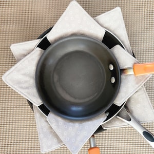 May include: Overhead view of a black frying pan with an orange handle, resting on a stack of two other pans. The pans are set on a beige patterned trivet. Kitchenware.