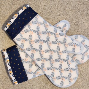May include: A pair of white oven mitts with a floral pattern. The mitts have a blue and yellow trim.
