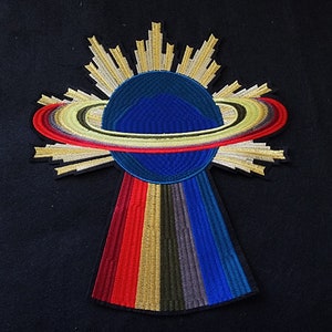 May include: A colorful embroidered patch depicting a planet with rings, surrounded by a sunburst design. The planet is blue with a green ring, and the sunburst is gold and white.