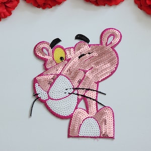 May include: A sequined iron-on patch featuring the Pink Panther character. The design is composed of pink and white sequins, with black thread detailing for the eyes, eyebrows, and whiskers. The character is shown in a winking pose.