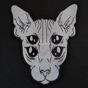 May include: Embroidered patch featuring a stylized cat face with four eyes. The patch is silver with black detailing and a black border. The cat's face has large, expressive eyes and a detailed nose and mouth.