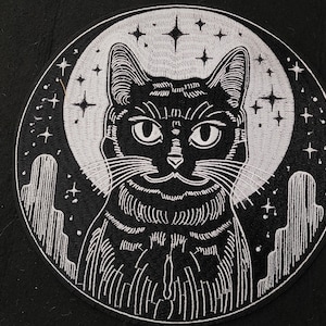 May include: Black and white embroidered patch of a cat with a crescent moon and stars in the background.