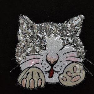 Peut inclure: Un visage de chat scintillant argenté et blanc avec une langue rose qui sort. Le chat cligne de l'œil et a une patte de chaque côté de son visage.