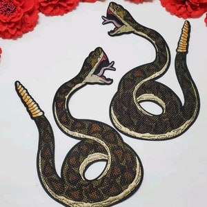 May include: Two embroidered iron-on patches of rattlesnakes with open mouths and detailed scales. The snakes are brown and black with a yellow underbelly and a red tongue.