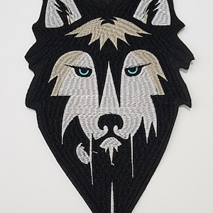 May include: Black and white embroidered wolf patch with teal eyes. The patch features a detailed design of a wolf's head, with intricate stitching and a diamond-shaped outline. The wolf's fur is depicted in shades of gray and black.
