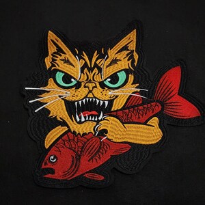 Peut inclure: Un patch brodé représentant un chat aux yeux verts et aux dents acérées tenant un poisson rouge dans ses pattes. Le chat est noir et orange.