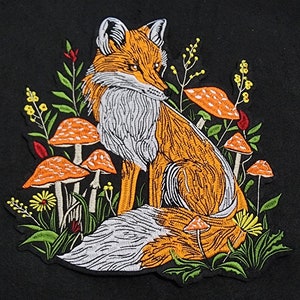 May include: Embroidered patch featuring a fox sitting amongst mushrooms and greenery. The fox is orange and white, with a bushy tail. The mushrooms are red and white with white stems. The greenery is green and yellow.