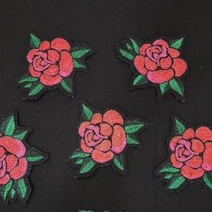 May include: Five embroidered rose patches with red petals and green leaves on a black background. The roses have a pink centre and are surrounded by green leaves. These patches can be used to decorate clothing or accessories.