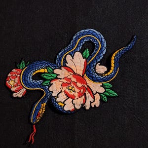 May include: Embroidered patch featuring a blue snake intertwined with a large peony flower. The snake has yellow and blue scales, a red tongue, and is surrounded by green leaves. The flower is pink and red. The patch is on a black background.