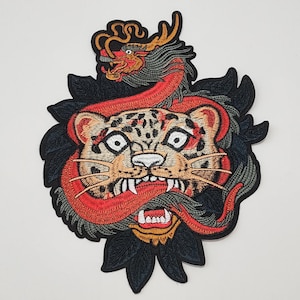 May include: Embroidered patch depicting a tiger and dragon design. The tiger has a tan face with black spots, white eyes, and a red mouth. The dragon is red and grey. Black leaves surround the tiger's head.