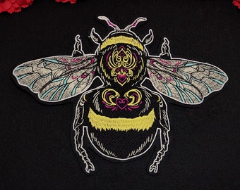 Peut inclure: Un écusson brodé représentant un bourdon stylisé avec un corps noir, une section médiane jaune duveteuse et des lignes décoratives complexes roses et jaunes. Les ailes sont détaillées avec des coutures bleu clair et roses sur fond sombre.