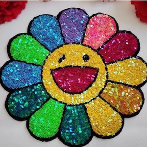 May include: A large, colorful, sequined flower patch with a smiling face in the center. The flower is made of iridescent sequins in shades of green, blue, purple, pink, and yellow.