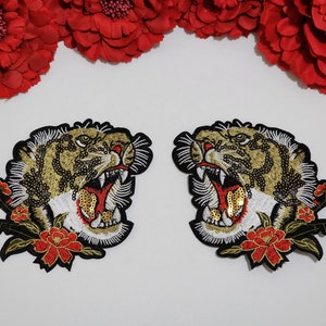 May include: Two embroidered tiger head patches featuring gold, black, and white thread with sequin accents. Each tiger has an open mouth showing white teeth and a red tongue, surrounded by red floral details with green leaves on a black border.