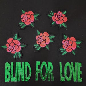 May include: Five embroidered patches featuring red and pink roses with green leaves. The patches are arranged on a black background, with the words "BLIND FOR LOVE" in green below the roses.