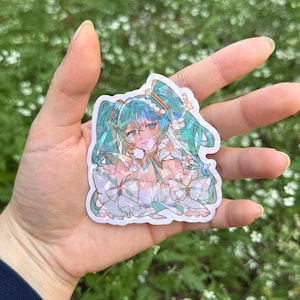 May include: A die-cut holographic sticker featuring an anime-style character with long teal twin-tails, wearing a white ruffled garment with large bows, adorned with white floral accents and a pearl-like necklace, rendered in a shimmering finish.