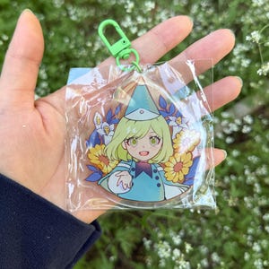May include: A circular acrylic keychain featuring an anime-style character with light green hair wearing a blue wizard hat and matching cape, surrounded by yellow sunflowers and blue leaves, attached to a bright green plastic lobster clasp.