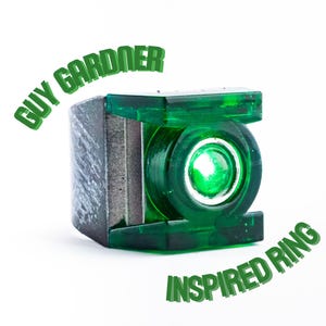 May include: A ring featuring a dark, textured metallic band and a translucent green rectangular face with a circular center. The text GUY GARDNER INSPIRED RING is arched around the object in green, blocky, three-dimensional lettering.