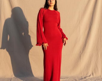 Puede incluir: Un vestido largo de punto rojo con mangas largas, que presenta un cuerpo y una falda ajustados. Las mangas son largas y anchas, terminando en un detalle de puño con volantes. El dobladillo del vestido también cuenta con un borde de volantes a juego.