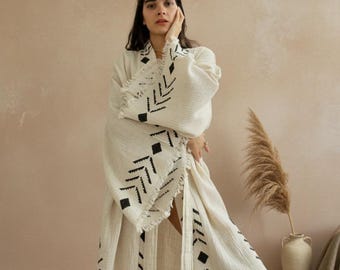May include: A long, cream-colored robe featuring a textured fabric and black geometric patterns along the front panels and wide sleeves. The garment includes frayed edges and a wrap-style design with a long, flowing silhouette.
