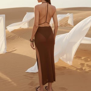 May include: A dark brown, floor-length skirt featuring a high side slit and a matching halter-style top with thin straps that cross over the open back. The garment is made of a fine-knit fabric and is shown from the back view in a desert setting.