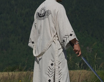 May include: A light-colored, long robe with black geometric patterns and a sun design on the back. The robe is worn with a belt and is paired with black pants. The person is holding a sword in a grassy field.