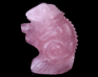 May include: A pink rose quartz carving of a chameleon. The chameleon is sitting on a branch with its tail curled around it. The carving is detailed and intricate.