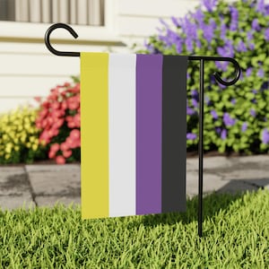 May include: A vertical garden flag featuring the non-binary pride flag design. The flag has five vertical stripes in yellow, white, purple, and black. The flag is displayed on a black metal garden stake, set in a grassy yard with flowering bushes in the background.