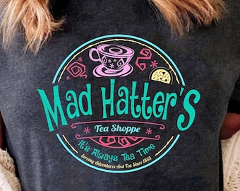 May include: A dark grey t-shirt with a teal circle graphic that says "Mad Hatter's Tea Shoppe" with a pink teacup and saucer, a lemon slice, and the text "It's Always Tea Time" and "Serving Adventures and Tea Since 1865".