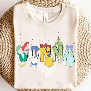 May include: A white t-shirt featuring a colourful design with Disney princesses and the name "JONNA". The princesses are dressed in their signature colours and are surrounded by stars and glitter.
