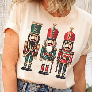 May include: A beige t-shirt featuring a graphic of three nutcracker soldiers. The nutcrackers are wearing red, green and blue uniforms with gold accents.