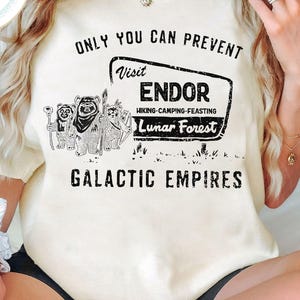Puede incluir: Una camiseta blanca con un letrero de estilo vintage que dice "Visita Endor - Senderismo-Camping-Festín - Bosque lunar" y el texto "Solo tú puedes prevenir los imperios galácticos". El letrero presenta una imagen de dibujos animados de cuatro Ewoks.