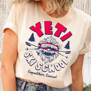 Puede incluir: Una camiseta de color crema con un estampado gr&aacute;fico de un yeti con gafas de esqu&iacute; y el texto "YETI SKI SCHOOL Expedition Everest".