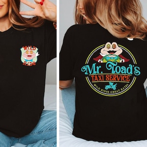 May include: Black t-shirt with a cartoon frog wearing a red jacket and driving a car. The text on the shirt says "Mr. Toad's Taxi Service" with the words "Wild Rides Great Rates" below the text.