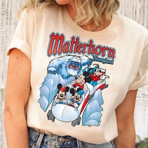 May include: A cream colored t-shirt with a graphic of Mickey Mouse and friends riding the Matterhorn ride at Disneyland. The text "Matterhorn Disneyland" is printed on the shirt.