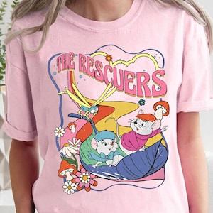 Op de afbeelding: Roze T-shirt met een grafische print van de Disney-personages uit De Reddertjes. De afbeelding toont de muizen Bernard en Miss Bianca, samen met een libel, en de tekst "The Rescuers".