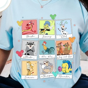 Puede incluir: Camiseta azul claro con una cuadrícula de retratos de personajes de dibujos animados, incluyendo Mushu, Jiminy, Olaf, Meeko, Hei Hei, Jag y Gus, Lumiere, Tambor y Flounder. La camiseta tiene cuello redondo y mangas cortas.