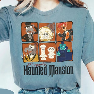 Può includere: Una maglietta blu con una grafica in stile vintage dei personaggi dei Muppet dell'attrazione Haunted Mansion. La grafica presenta Gonzo, Rizzo, il Ghost Host e altri. Il testo "Muppets Haunted Mansion" è stampato sotto la grafica.