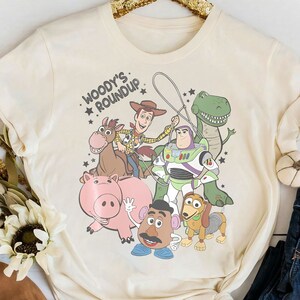 Puede incluir: Camiseta de color crema con un gr&aacute;fico de personajes de la pel&iacute;cula animada Toy Story. El dise&ntilde;o incluye a Woody, Buzz Lightyear, Rex, Hamm, Sr. Potato, Slinky Dog y Bullseye, con el texto "Woody's Roundup."