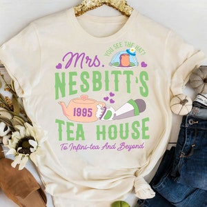 K&ouml;nnte beinhalten: Ein cremefarbenes T-Shirt mit einem gr&uuml;nen und violetten Grafikdesign, auf dem "Mrs. Nesbitt's Tea House" steht, mit einem Cartoon-Bild einer Teekanne und einer Figur aus dem Film Toy Story. Der Text lautet auch "You see the hat?" und "1995" und "To Infini-tea And Beyond".
