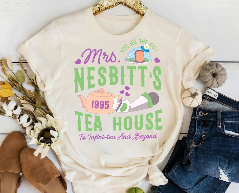 K&ouml;nnte beinhalten: Ein cremefarbenes T-Shirt mit einem gr&uuml;nen und violetten Grafikdesign, auf dem "Mrs. Nesbitt's Tea House" steht, mit einem Cartoon-Bild einer Teekanne und einer Figur aus dem Film Toy Story. Der Text lautet auch "You see the hat?" und "1995" und "To Infini-tea And Beyond".