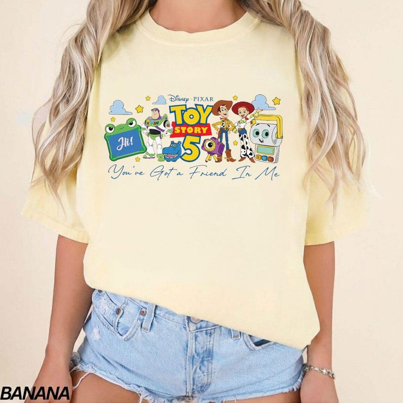 May include: A light yellow t-shirt featuring a graphic from the Disney Pixar movie Toy Story 5. The graphic includes the characters Woody, Buzz Lightyear, and others, with the text "Toy Story 5" and "You've Got a Friend In Me".