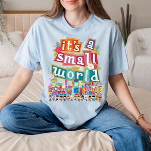 Puede incluir: Una camiseta azul claro con un estampado gr&aacute;fico colorido que dice "it's a small world" con una representaci&oacute;n del ic&oacute;nico paseo de Disney.