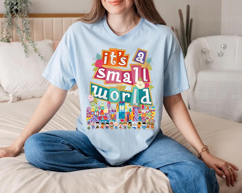 Puede incluir: Una camiseta azul claro con un estampado gr&aacute;fico colorido que dice "it's a small world" con una representaci&oacute;n del ic&oacute;nico paseo de Disney.