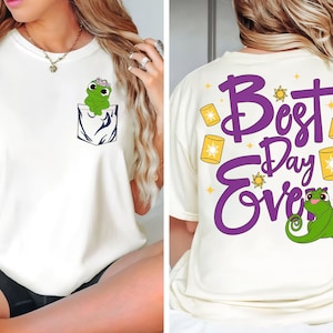 Op de afbeelding: Wit T-shirt met een groene kikker met een bloemenkroon in een zakje op de voorkant en paarse tekst die "Best Day Ever" zegt met een groene kameleon op de achterkant.