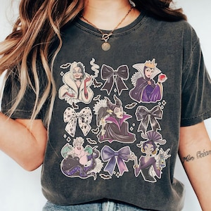 May include: Dark gray t-shirt featuring illustrations of cartoon villains and black bows. The graphic includes characters like Cruella de Vil, the Evil Queen, and Ursula. The design is surrounded by small bat and spiderweb graphics.
