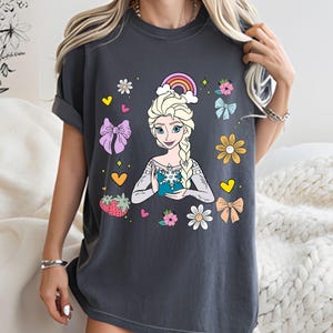 May include: A dark gray t-shirt with a colorful graphic of a cartoon princess holding a snowflake. The graphic includes a rainbow, hearts, flowers, and bows.